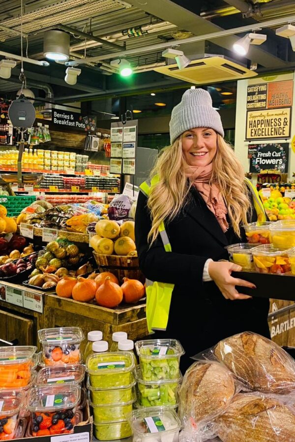A female volunteer holding a basket of food whilst in the fruit and veg section of Wholefoods