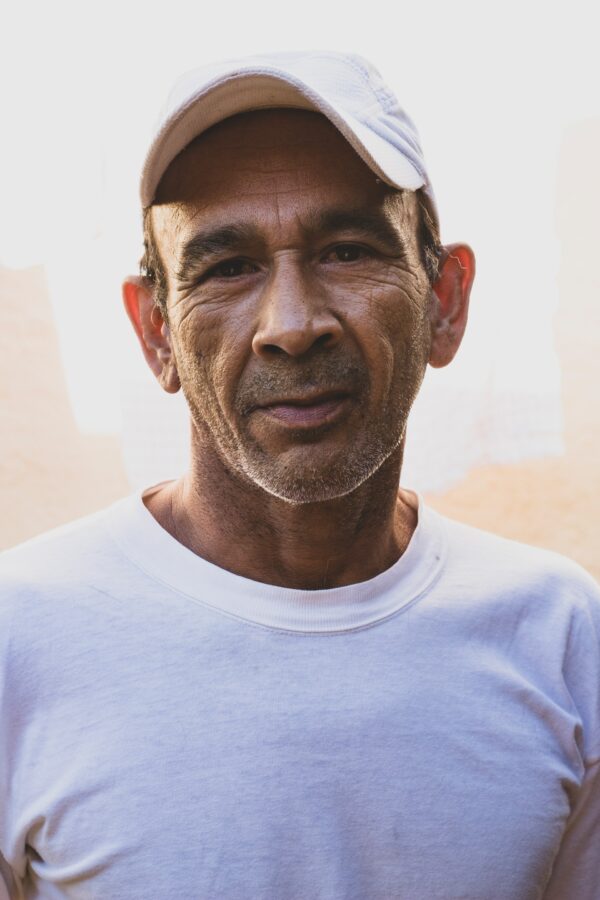 Older man with some stubble tanned and wearing a cap with a white t-shirt looking straight into the camera